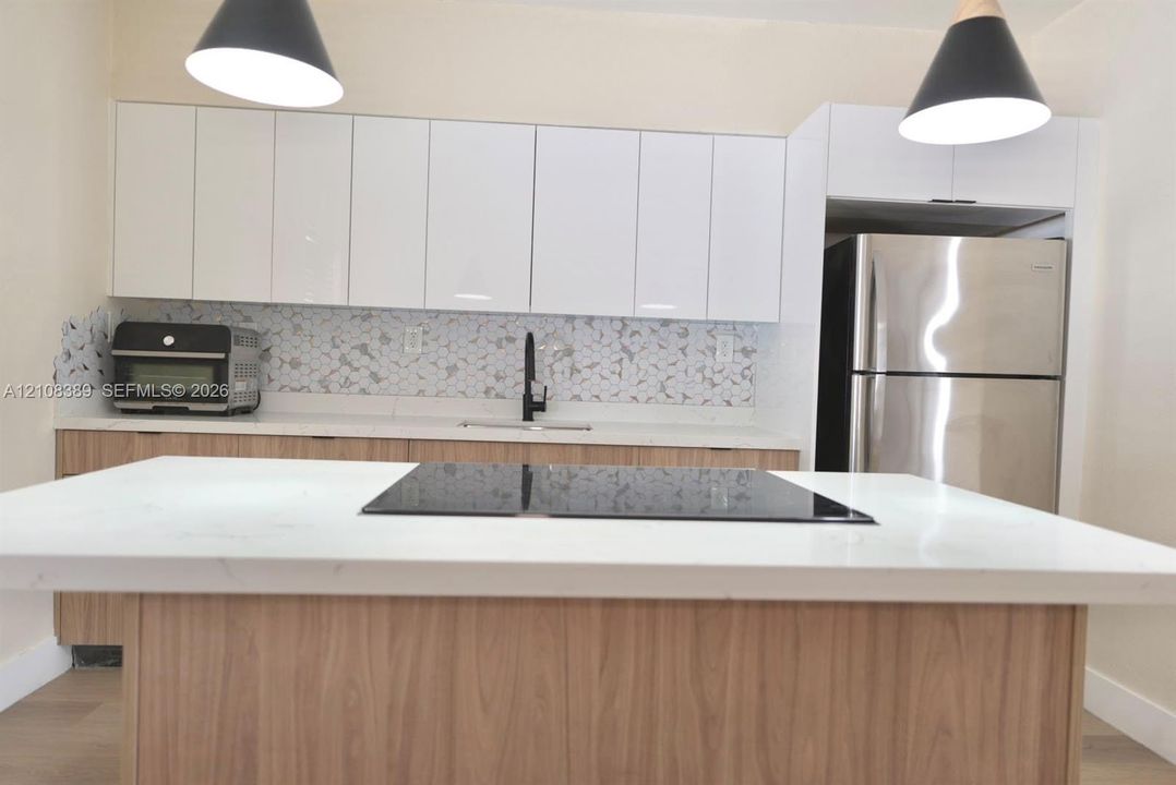 Kitchen featuring white high-gloss upper cabinetry, wood-finish lower cabinetry, stone-surface countertops, a center island with a built-in cooktop, and a patterned tile backsplash