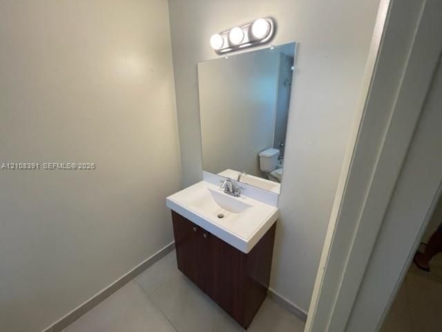 Vanity with integrated sink basin, dark wood-finish cabinetry, rectangular wall mirror, three-bulb vanity lighting fixture, and ceramic tile flooring