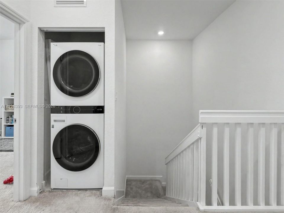 Stacked laundry unit with front-loading appliances, recessed ceiling lighting, and white wooden stair railing
