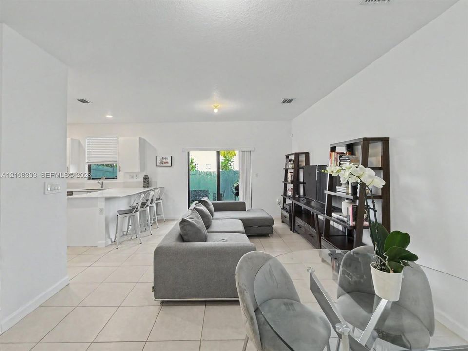 Open-concept interior featuring neutral ceramic tile flooring, solid-surface kitchen countertops, a center island with breakfast bar seating, and glass sliding doors leading to an exterior space