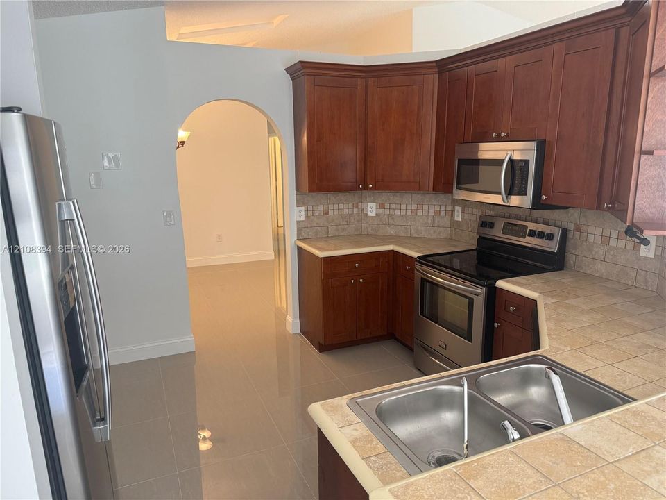 Kitchen featuring cherry-finish cabinetry, stainless steel appliances, tile countertops, a mosaic tile backsplash, and ceramic tile flooring