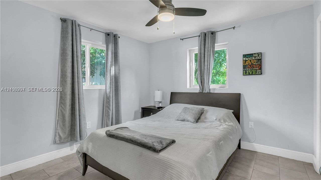 Neutral-toned bedroom featuring large format tile flooring, light-colored walls, dual windows, and a central ceiling fan with integrated lighting