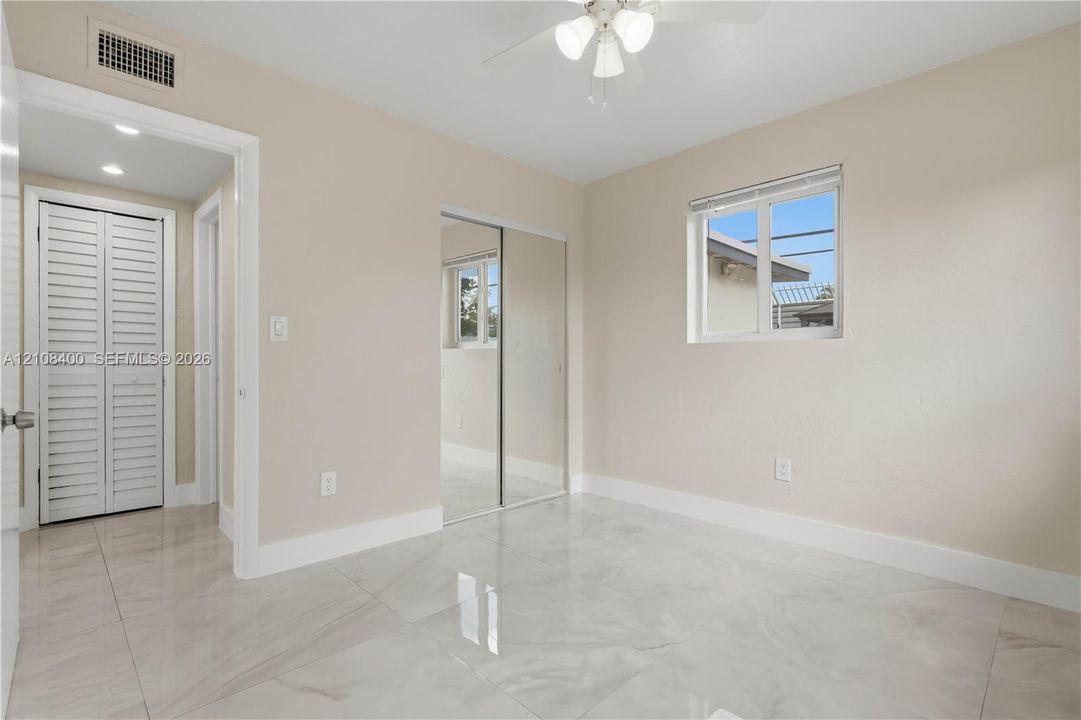 Neutral-toned room featuring large-format tile flooring, mirrored sliding-door closet, multi-pane window, and integrated ceiling fan