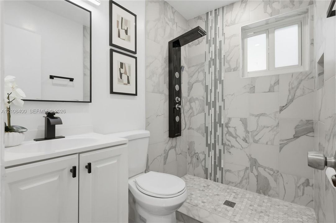 Bathroom featuring large-format wall tile, shower panel system with integrated body jets, walk-in shower with pebble tile flooring, vanity with matte black faucet hardware, and a framed mirror