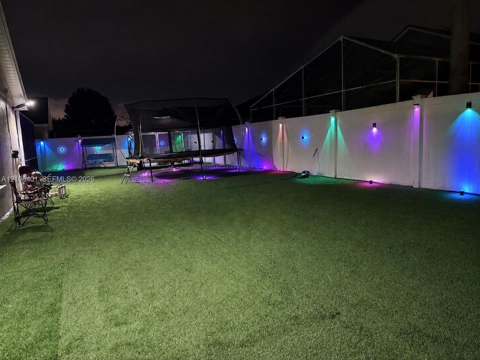Fenced backyard featuring synthetic turf, privacy screening, and wall-mounted exterior accent lighting