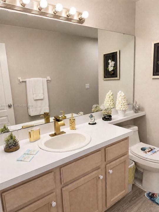 Bathroom vanity featuring a white countertop, inset oval sink, gold-toned faucet, wood-finish cabinetry, and a full-width wall mirror with integrated lighting