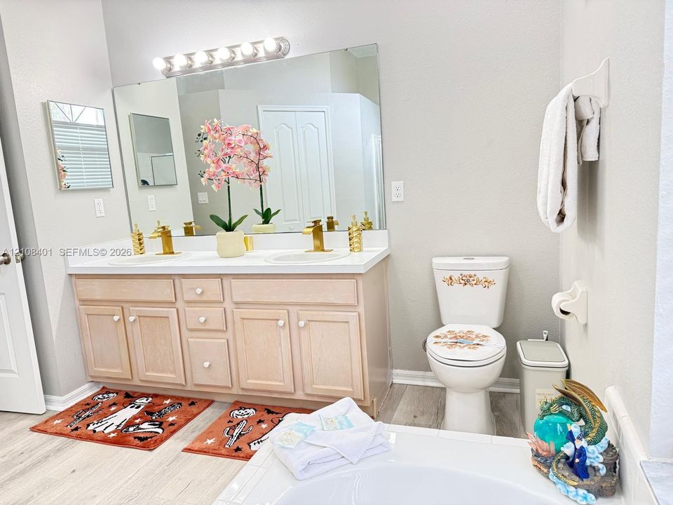 Dual-vanity cabinetry with light wood-finish, white solid-surface countertop, gold-toned faucets, full-width wall mirror, and integrated bar lighting