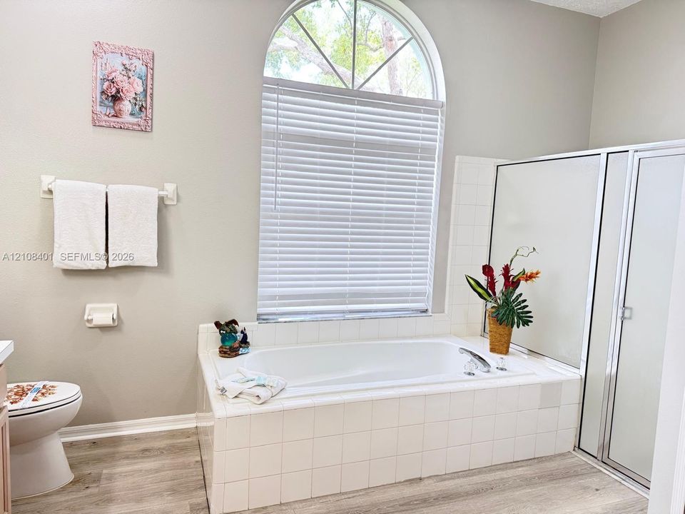 Built-in soaking tub with a tiled surround, arched window with a muntin grid, walk-in shower with a framed glass enclosure, and wood-finish flooring