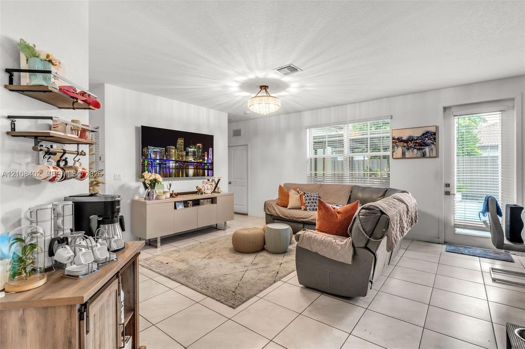 Living area featuring tile flooring, a multi-tiered crystal-style chandelier, horizontal window blinds, and a glass-paneled exterior door