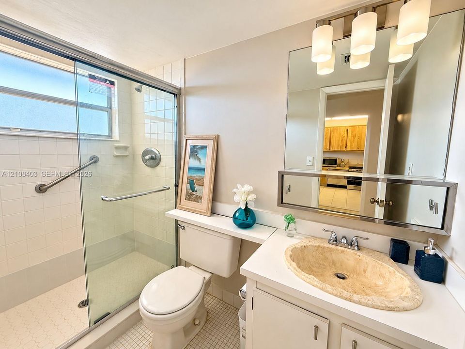 Inviting bathroom featuring a glass-enclosed shower, offering a clean, modern look with tile surround, vanity with a stone-finish oval sink, and a large wall-mounted mirror with integrated lighting