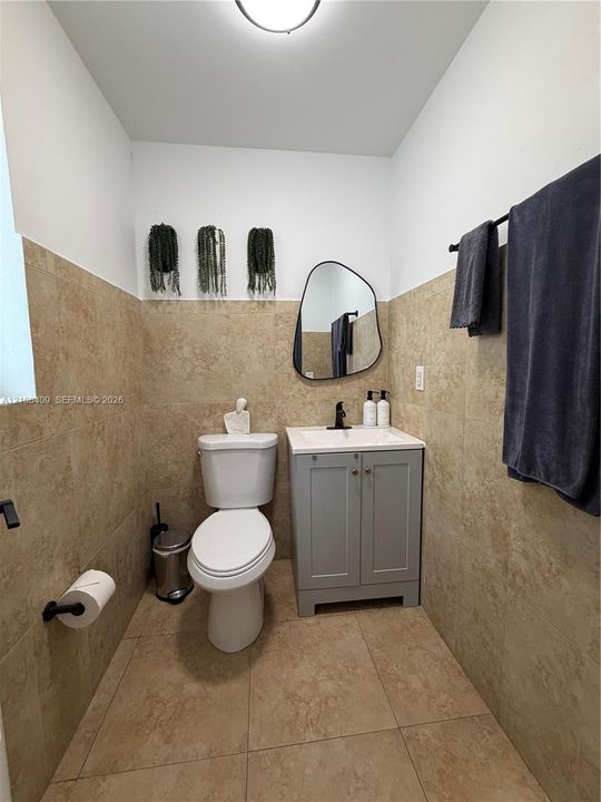 Neutral-toned wall and floor tiling, gray vanity cabinet with a white countertop, organic-shaped wall mirror, and matte black hardware