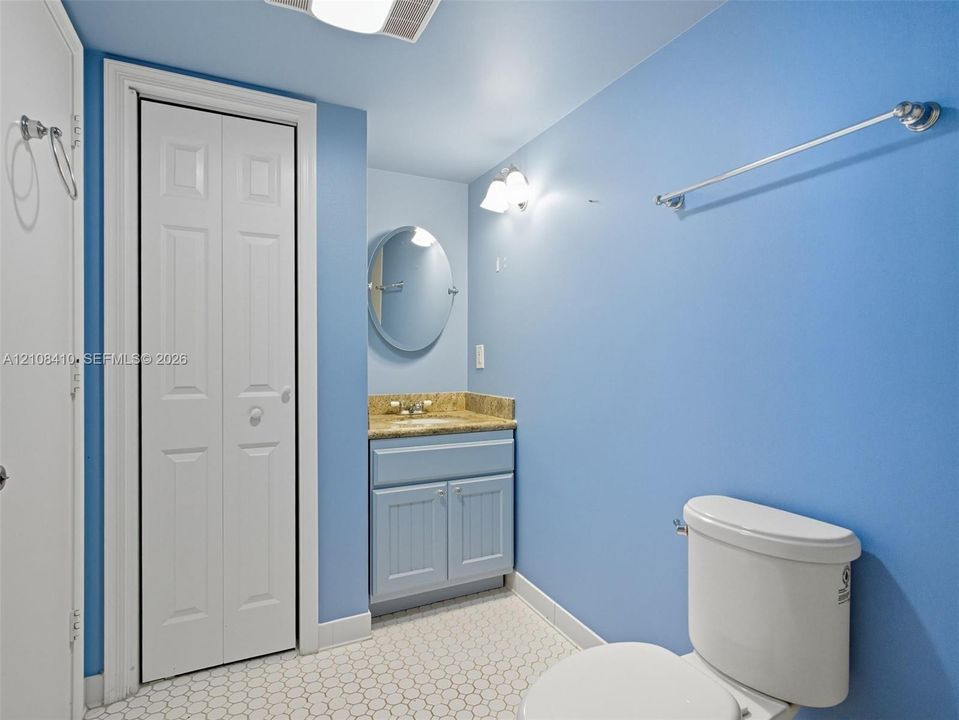 Bathroom featuring a single vanity with stone-style countertop, oval wall-mounted mirror, white ceramic tile flooring, bifold closet doors, and brushed metallic hardware