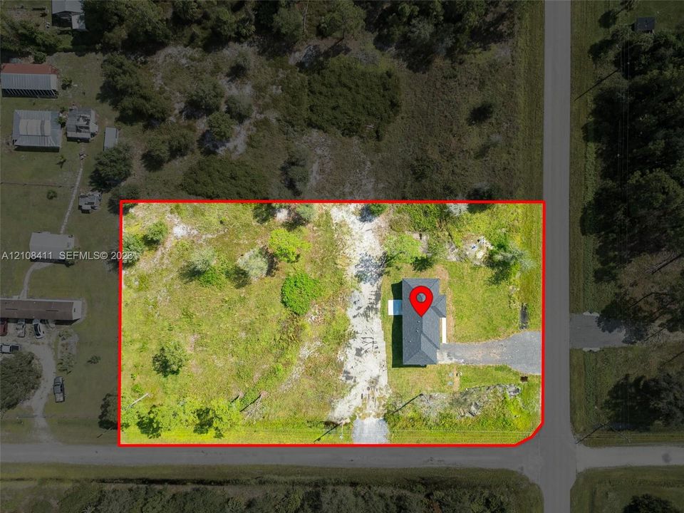 Aerial view of a corner lot featuring a detached structure, gravel driveway, mature trees, and expansive open green space