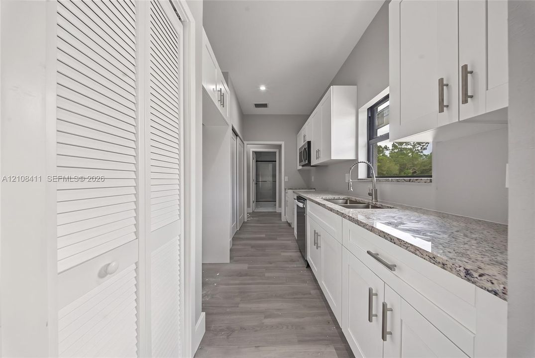 Galley kitchen featuring white Shaker-style cabinetry, stone-finish countertops, stainless steel undermount sink with a gooseneck faucet, wood-finish flooring, and white louvered-door closets