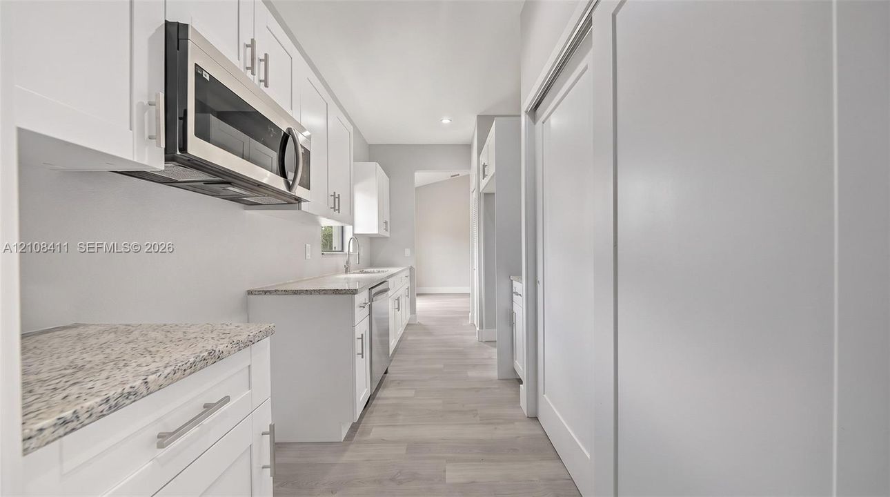 White shaker-style cabinetry featuring satin nickel hardware, speckled stone-finish countertops, stainless steel over-the-range microwave, built-in dishwasher, and wood-finish flooring