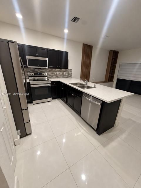 Kitchen featuring dark cabinetry, stone-finish countertops, stainless steel appliances, a multi-tone tile backsplash, and oversized light-colored floor tiling