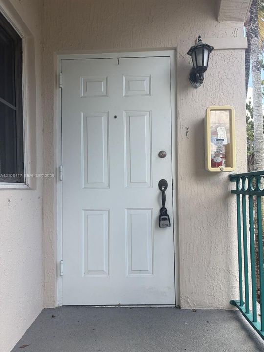 Six-panel entry door featuring a matte black handle set and peephole, complemented by an exterior wall-mounted carriage-style lantern and a recessed metal wall cabinet