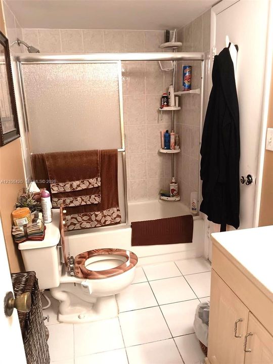 Bathroom featuring a tub-shower combination with a sliding glass door enclosure and light-toned tile surround, white square floor tiling, and wood-finish cabinetry