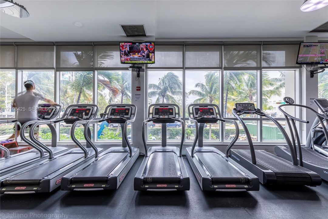 Fitness center featuring floor-to-ceiling windows with roller shades, recessed lighting, and integrated wall-mounted televisions