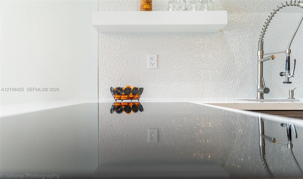 Polished solid-surface countertop with an integrated cooktop, textured white tile backsplash, built-in floating shelf, and stainless steel high-arc pull-down faucet