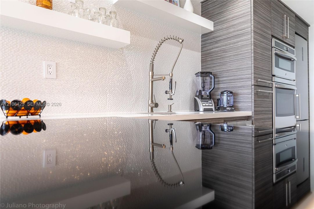 Textured white backsplash, polished dark countertop, professional-style pull-down faucet, built-in floating shelf, and stacked stainless steel oven and microwave