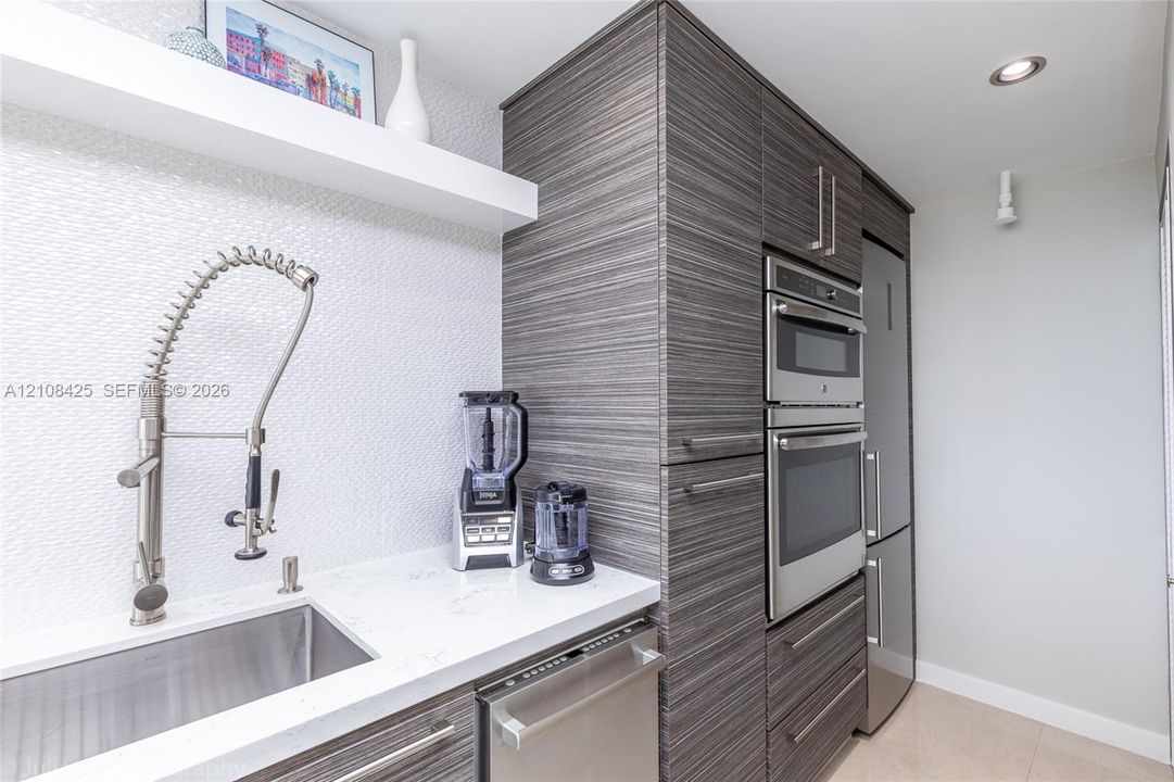 Kitchen featuring textured cabinetry, stone-finish countertops, stainless steel appliances, a pull-down sprayer faucet, and a wall-mounted floating shelf