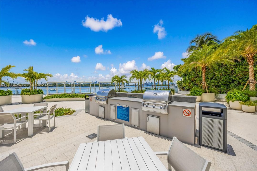 Outdoor kitchen featuring a built-in stainless steel grill, stone countertop, integrated cabinetry, paved patio flooring, and mature palm landscaping