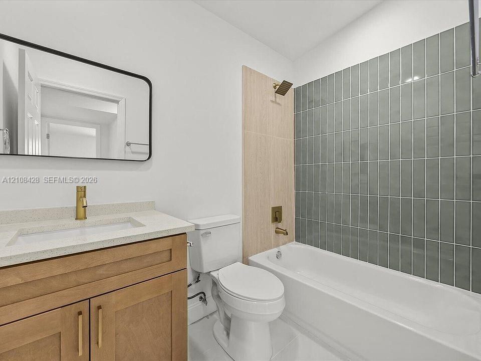 Bathroom featuring a wood-finish vanity with a stone-composite countertop, rectangular undermount sink, brass-finish fixtures, and a glass-tiled shower surround