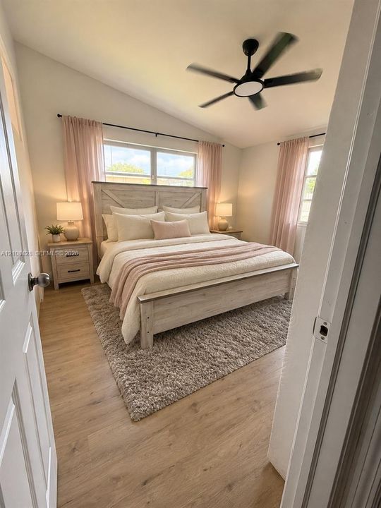 Vaulted ceilings, wood-finish flooring, dual windows with black curtain rods, and a multi-blade ceiling fan with integrated lighting