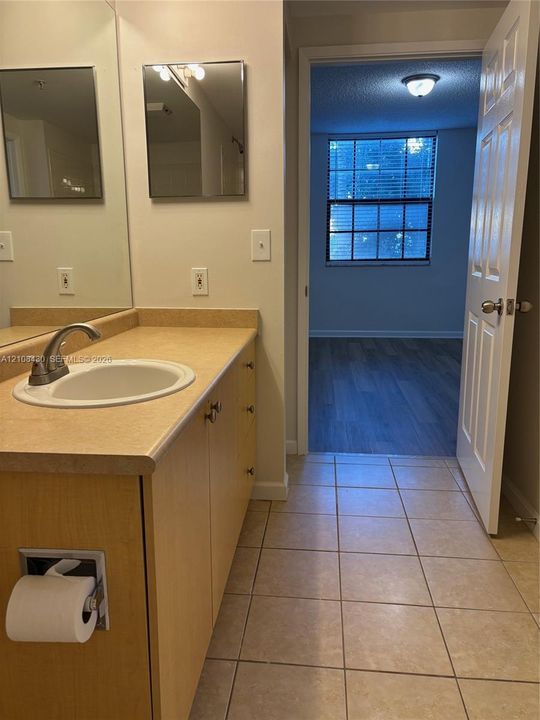 Bathroom featuring a laminate countertop, integrated sink with a curved faucet, light wood-finish cabinetry, ceramic tile flooring, and a flat-panel mirror