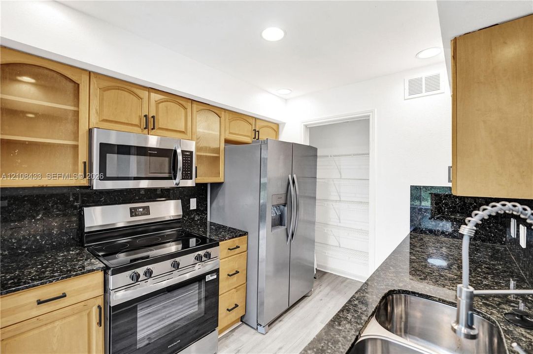 Kitchen featuring light-finish cabinetry with glass-front accents, dark stone countertops, stainless steel appliances, dual-basin stainless steel sink with pull-down faucet, and walk-in pantry