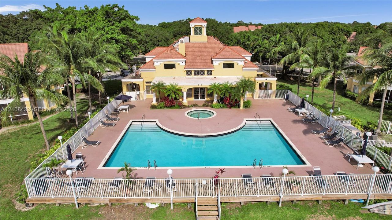Mediterranean-style clubhouse featuring a terra-cotta tile roof and a central observation tower, accompanied by a fenced outdoor pool and spa surrounded by a paved deck