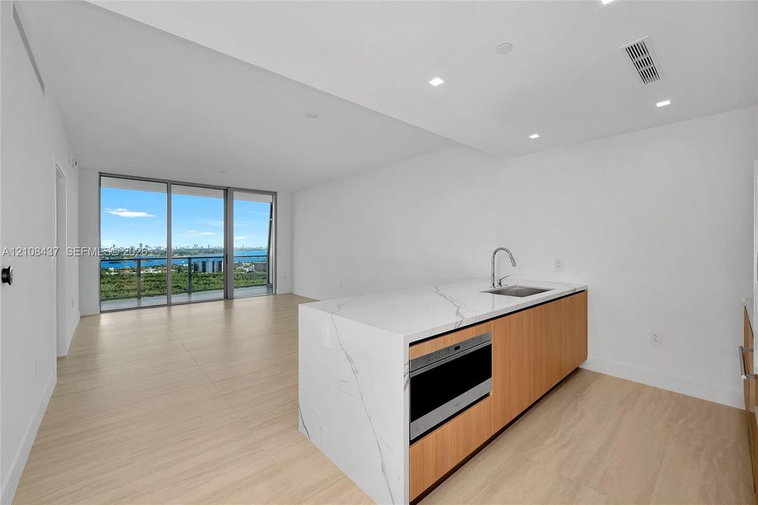 Kitchen island featuring stone-surfaced waterfall countertop, wood-finish cabinetry, stainless steel sink with high-arch faucet, and built-in microwave, complemented by wood-finish flooring and floor-to-ceiling glass sliding doors