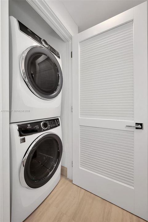 Built-in stacked laundry appliance set with wood-finish flooring and a white louvered door featuring polished chrome hardware