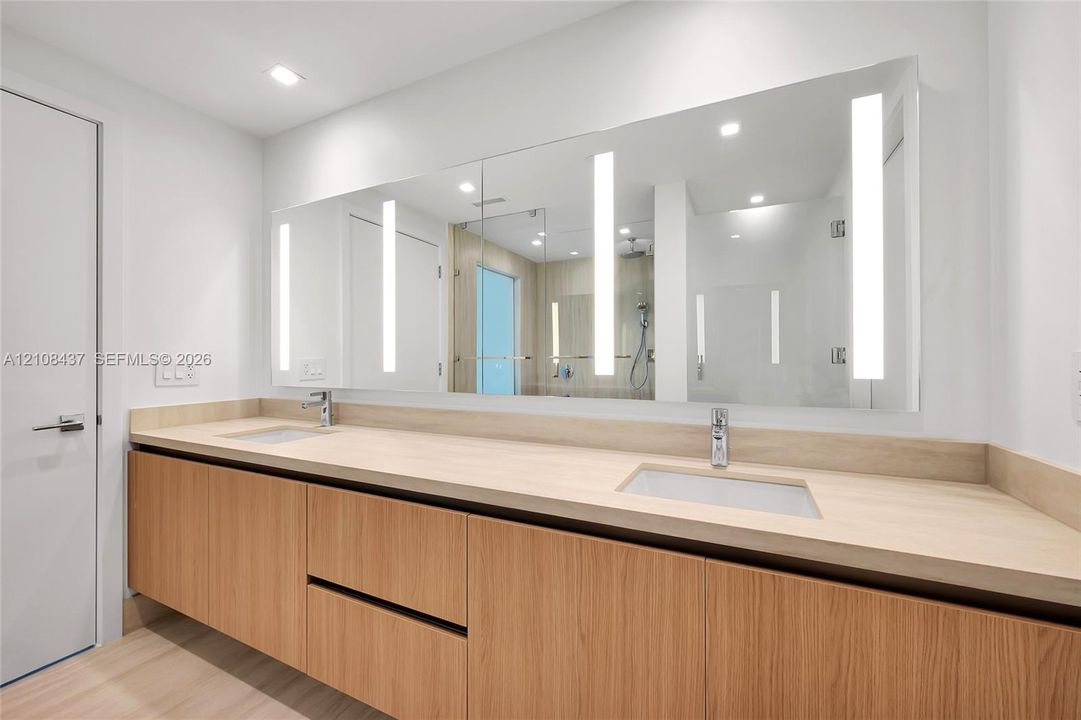Floating dual-sink vanity featuring wood-finish cabinetry, a light-toned solid surface countertop, rectangular undermount sinks, and a full-width integrated LED vanity mirror