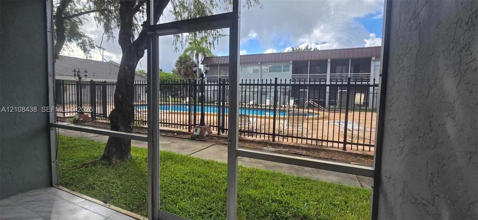 Screened-in patio with tile flooring, featuring proximity to a community swimming pool enclosed by black metal fencing and mature landscaping