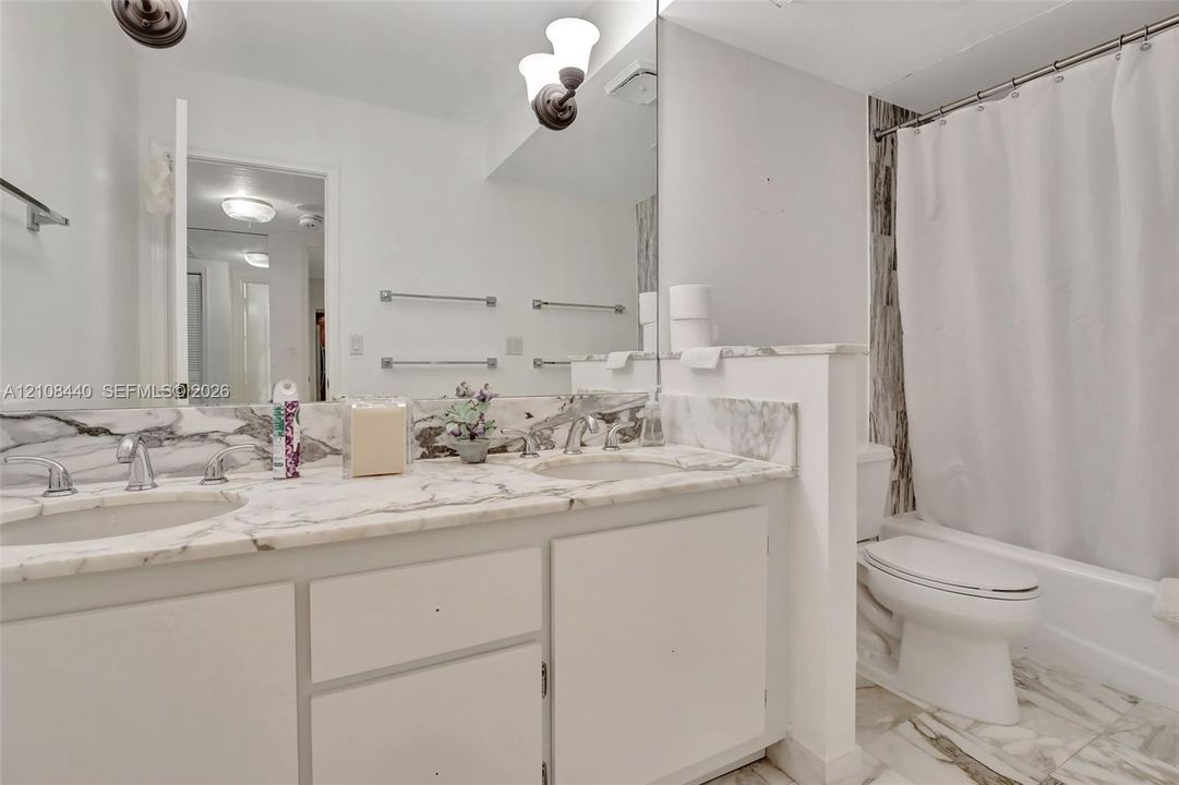 Dual-sink vanity with stone-finish countertop and backsplash, wall-to-wall mirror, brushed metal hardware, and tub-shower combination with floor-to-ceiling stone-finish surround