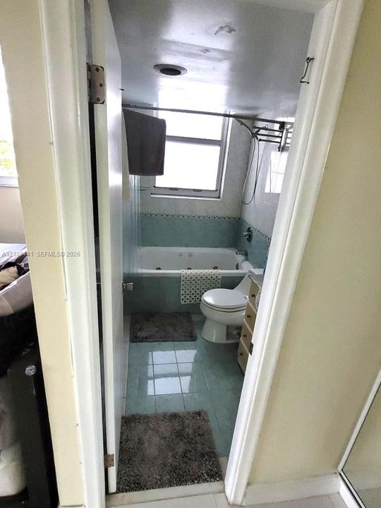 Jetted soaking tub with tile surround, porcelain toilet, and light-toned tile flooring