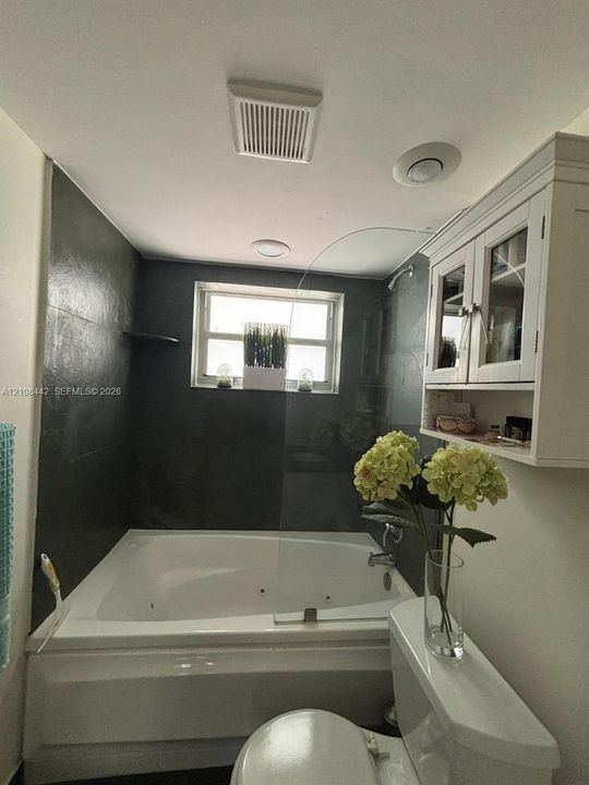 Jetted soaking tub with integrated shower hardware, dark tile surround, fixed glass partition, and wall-mounted white cabinetry