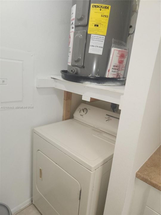 Utility closet featuring an electric water heater housed on a support shelf above a clothes dryer