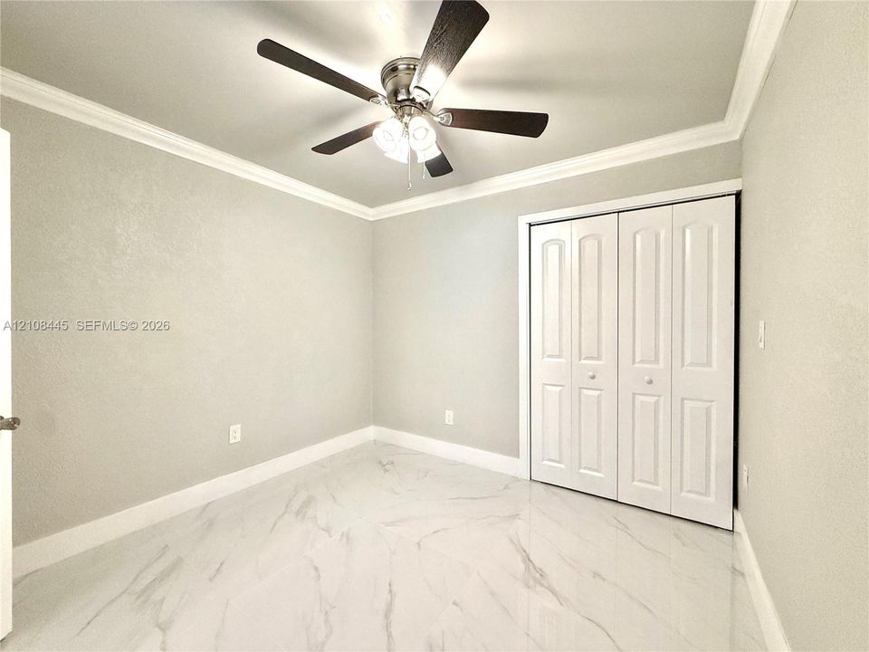 Downstairs Bedroom. Large-format tile flooring with a neutral veined pattern, crown molding, ceiling fan with integrated lighting, and white bi-fold closet doors