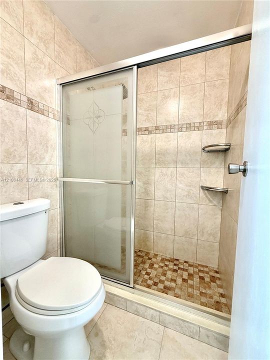 Shower enclosure featuring a sliding glass door with frosted detailing, neutral-toned tiled wall surrounds with decorative mosaic accents, integrated shower shelving, and a patterned tiled shower floor