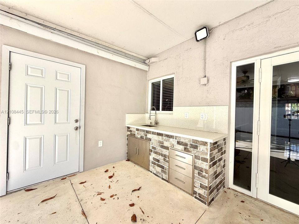 Covered outdoor kitchen area featuring a stone-veneer base, solid surface countertop, stainless steel integrated sink, and built-in metal cabinetry