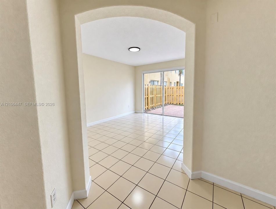 Arched entryway leading to a room with neutral ceramic tile flooring, flush-mount lighting, and glass sliding doors opening to a paved outdoor area with wood-style fencing