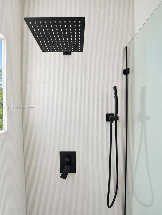 Shower enclosure featuring textured vertical-groove wall tiling, a square overhead rainfall shower head, a separate handheld sprayer, and matte black fixtures protected by a glass panel