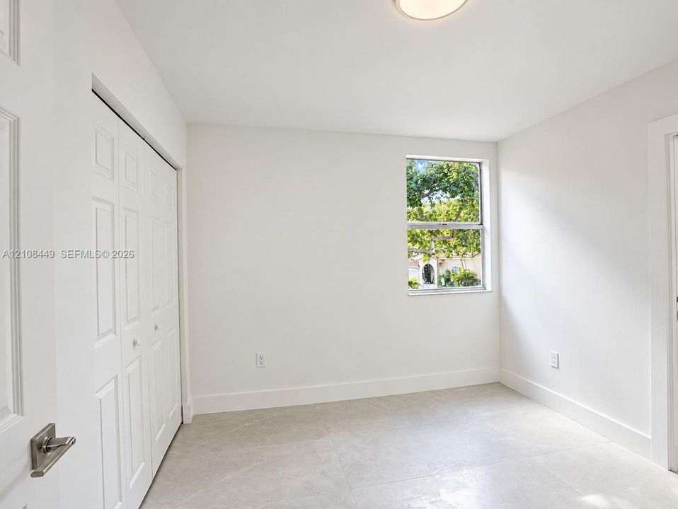 Neutral interior space featuring large-format tile flooring, a multi-panel bi-fold closet, a single-hung window with exterior greenery, and a flush-mount ceiling light fixture