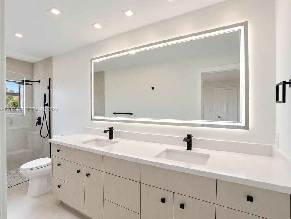 Double vanity featuring wood-finish cabinetry, solid-surface countertops, integrated dual sinks, and matte black hardware, complemented by a large LED-backlit mirror and a walk-in glass shower enclosure