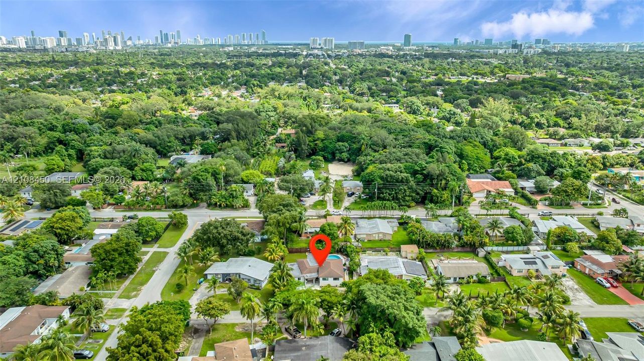 Aerial perspective featuring a property with a swimming pool, situated within a landscape of mature trees and established residential vegetation, incorporating a backdrop of an urban skyline