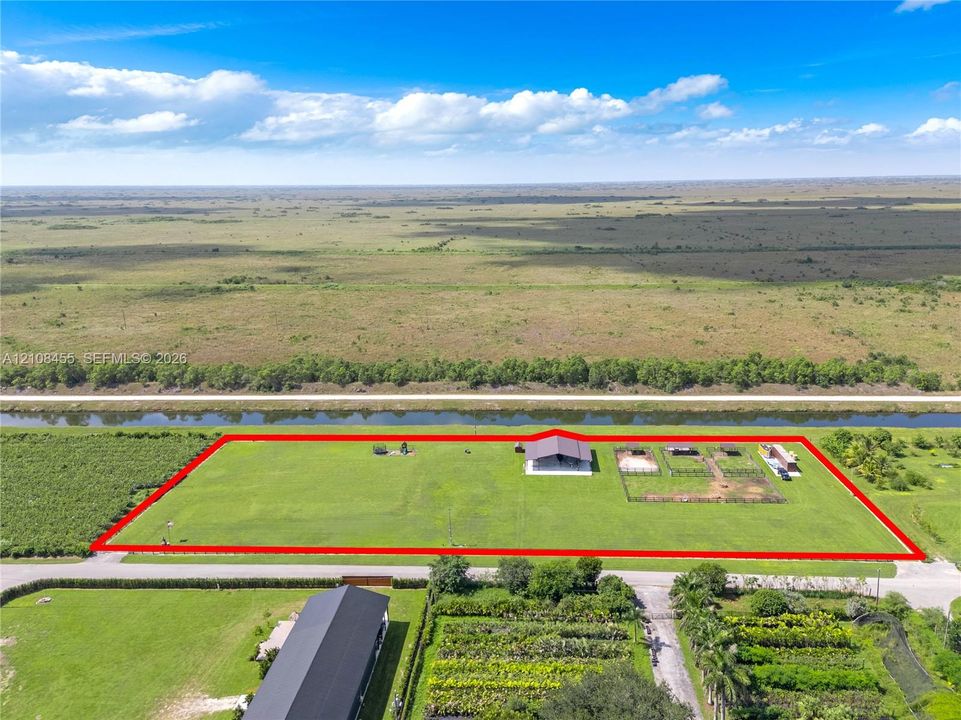Expansive acreage featuring a metal-roof structure, fenced outdoor enclosures, a canal-front perimeter, and open grassland