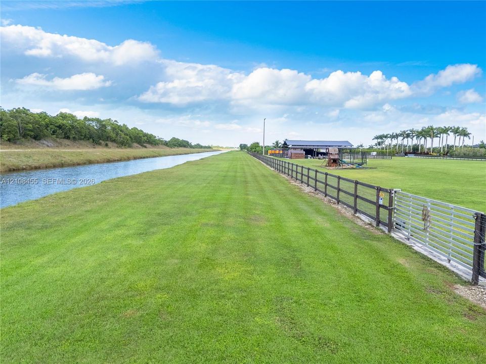 Expansive acreage featuring a canal-front perimeter, manicured pastureland, a perimeter post-and-rail fence, a metal gate, and an open-sided pole barn structure